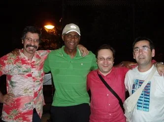 Four people standing outdoors at night, smiling at the camera. The first person is wearing a colorful Hawaiian shirt, the second person is in a green polo shirt and white cap, the third person is wearing glasses and a red shirt, and the fourth person