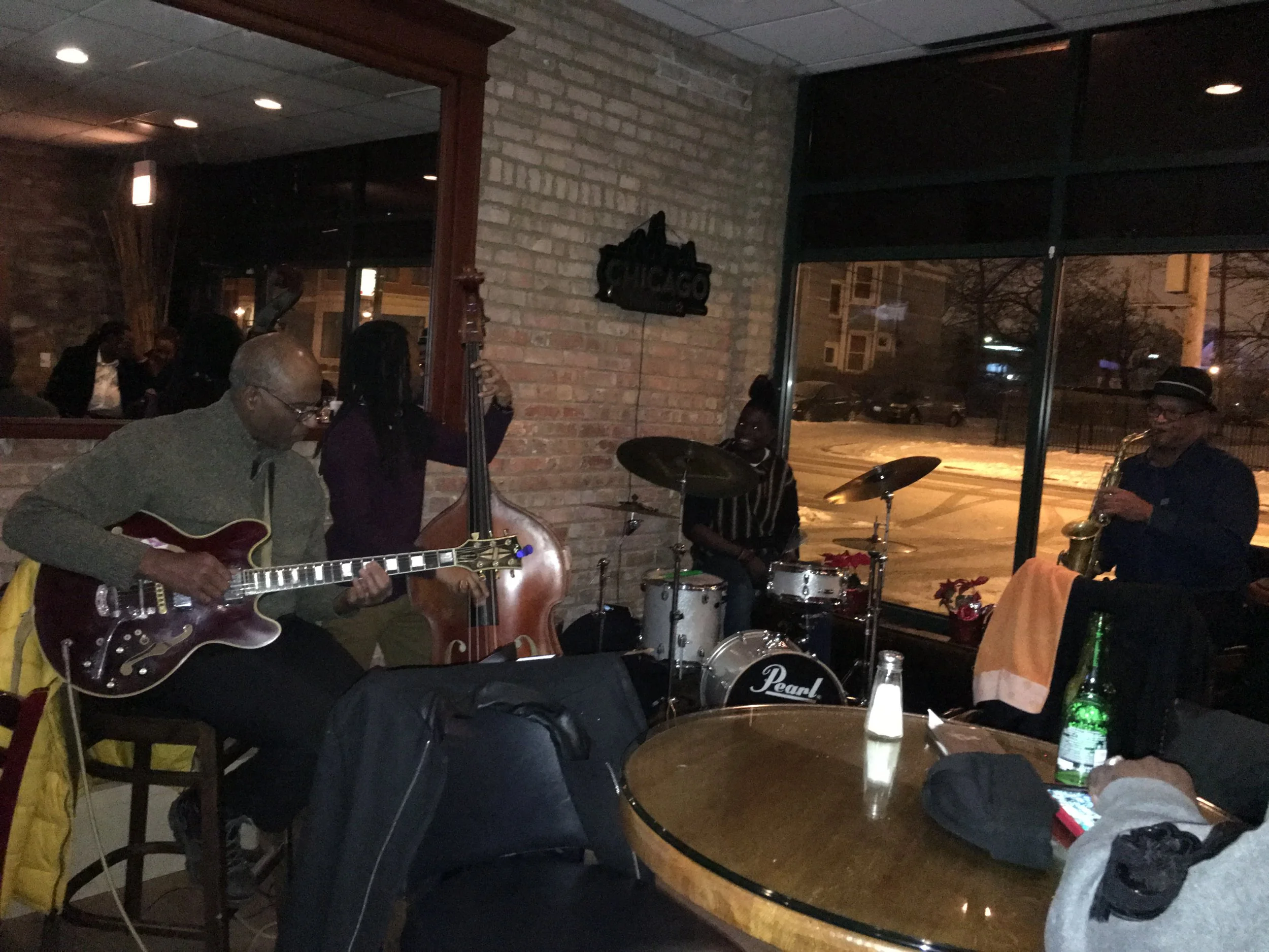A jazz band performing in a dimly lit restaurant or bar with brick walls. The band includes a guitarist, a double bassist, a drummer, and a saxophonist. Several patrons are seated at tables, some watching the performance and one using a smartphone. T