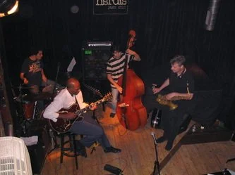 Four musicians playing jazz instruments on stage: one with drums, one with an acoustic guitar, one with an upright bass, and one with an electric guitar.