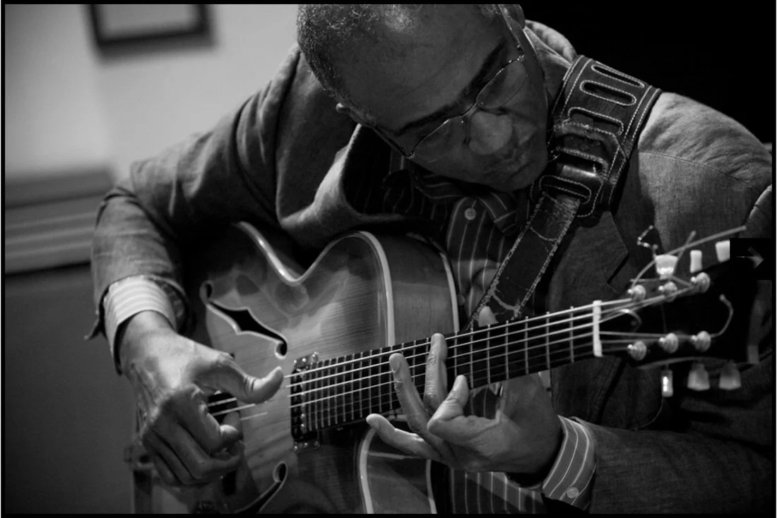 Black and white photo of a man playing an acoustic guitar, wearing glasses and a suit.