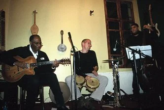 Three musicians performing in a room: a man playing guitar, a woman playing bongos, and another man at a keyboard.