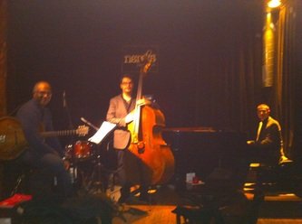 Three musicians playing jazz instruments on a dimly lit stage, including a guitarist, a double bassist, and a pianist.
