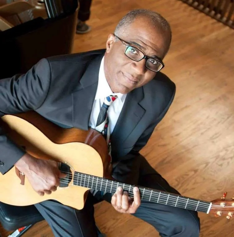 A man in a suit and glasses sitting on a wooden floor, holding and playing an acoustic guitar, looking up at the camera.