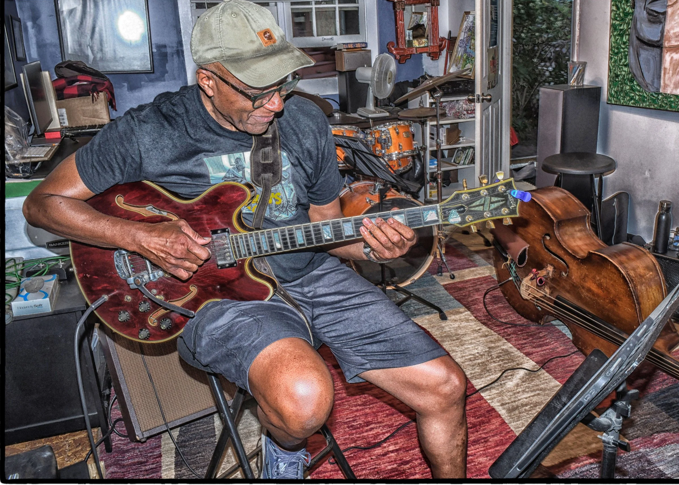 A man playing an electric guitar in a music studio, with a double bass lying on the floor nearby.
