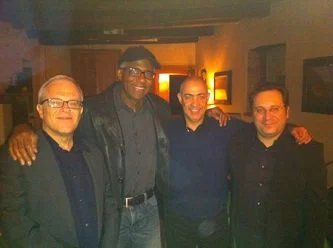 Four men standing together indoors, smiling at the camera with warm lighting in the background.