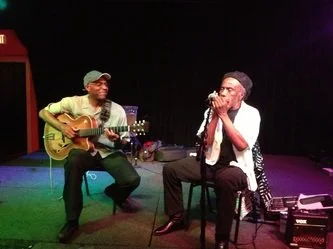 Two musicians performing on stage, one playing an acoustic guitar and the other singing into a microphone.