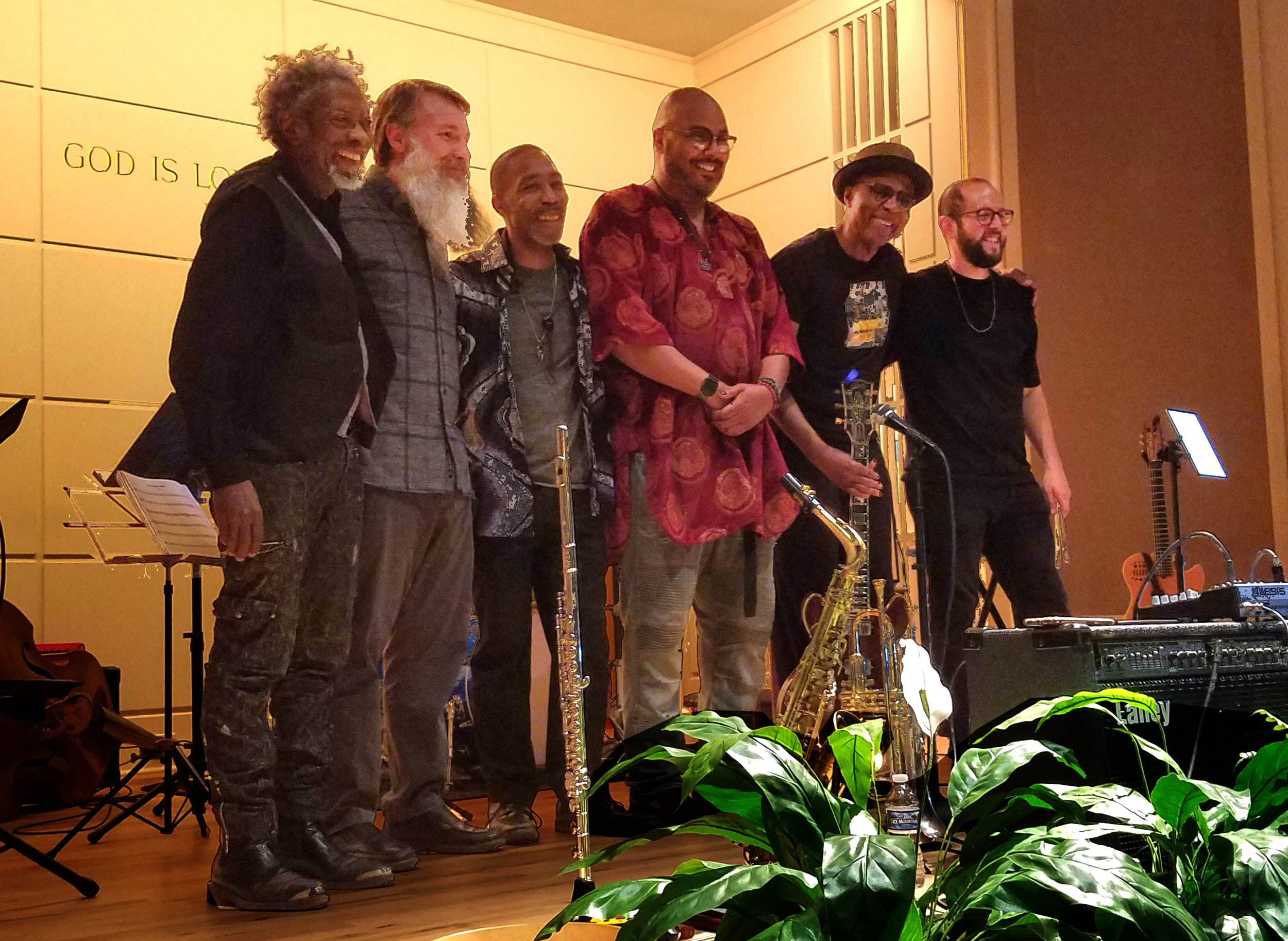 A group of seven musicians standing on stage, smiling. They have various musical instruments, including saxophones. There are plants in the foreground and music stands and guitars in the background.