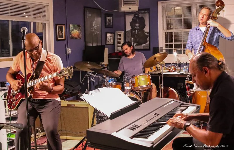 Four musicians playing instruments in a cozy indoor space, with paintings and photographs on the wall, including a photo of a man and a woman. The band includes a guitarist, a drummer, a double bass player, and a keyboardist.