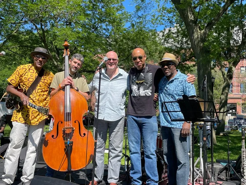 Group of five men standing outdoors on a sunny day, some holding musical instruments, with trees and buildings in the background.