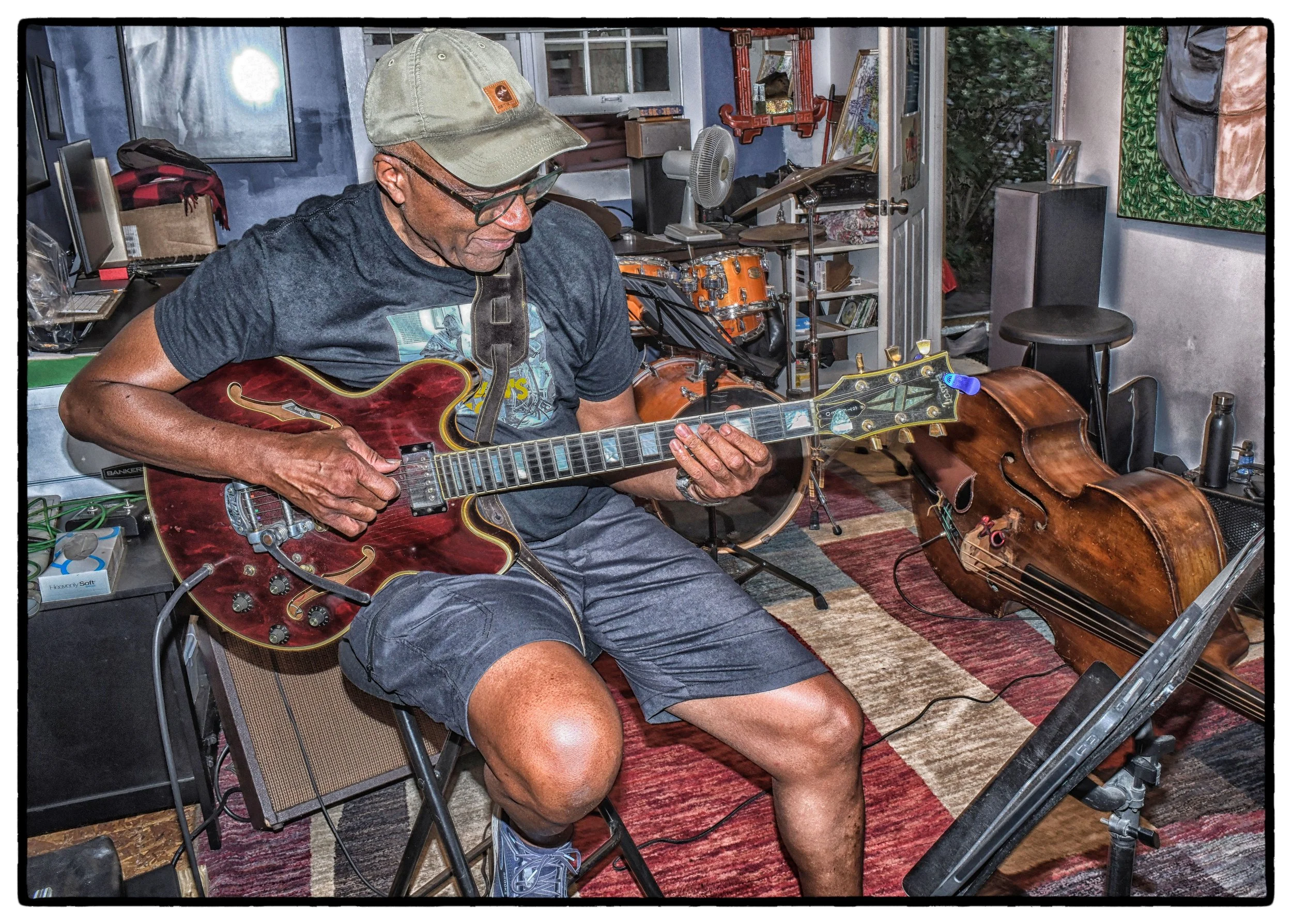 A man playing an electric guitar in a music studio with a cello on a stand next to him.
