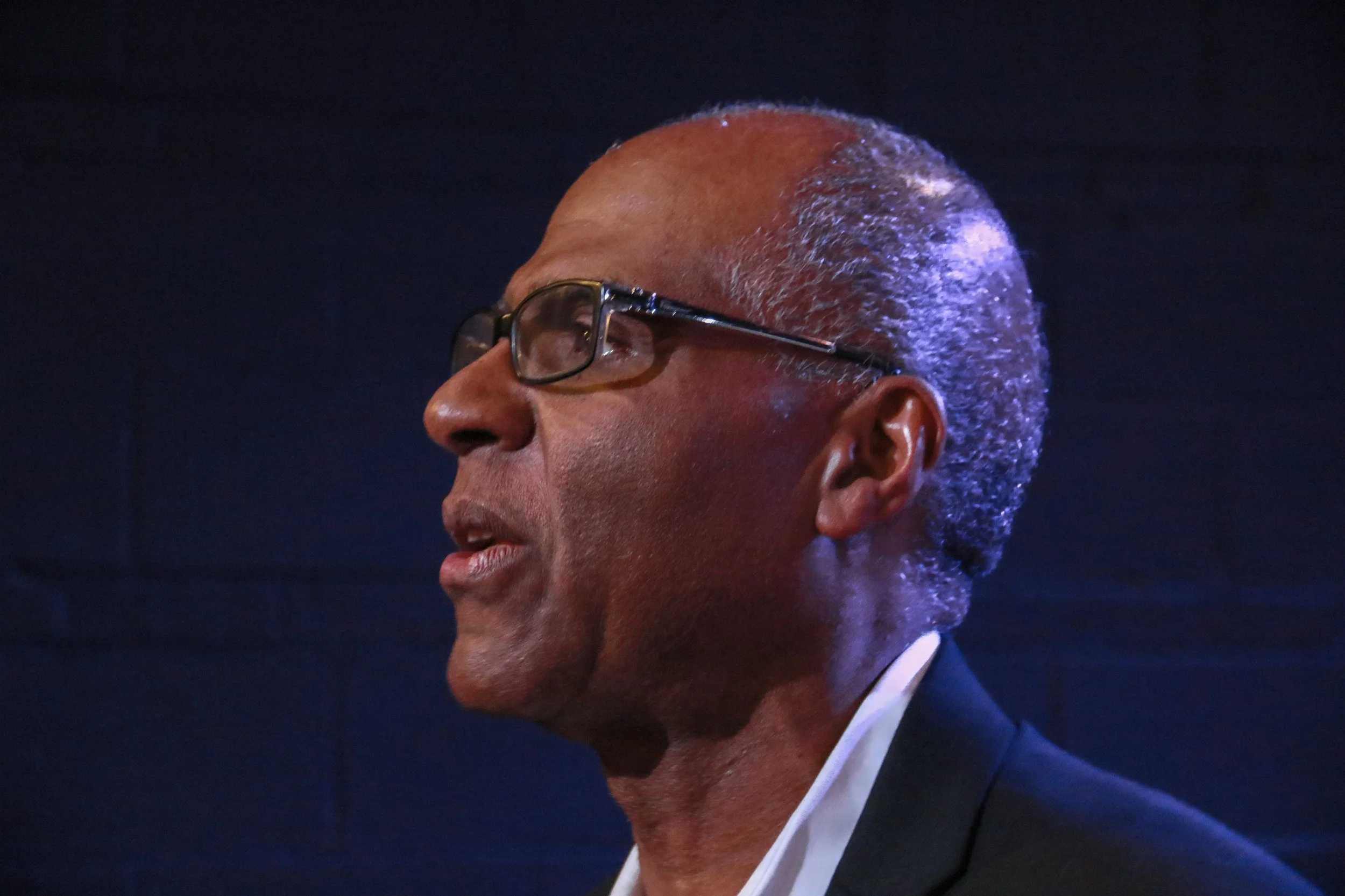 A profile view of an elderly man wearing glasses, speaking or listening, with gray hair, dark suit, and a light shirt against a dark background.