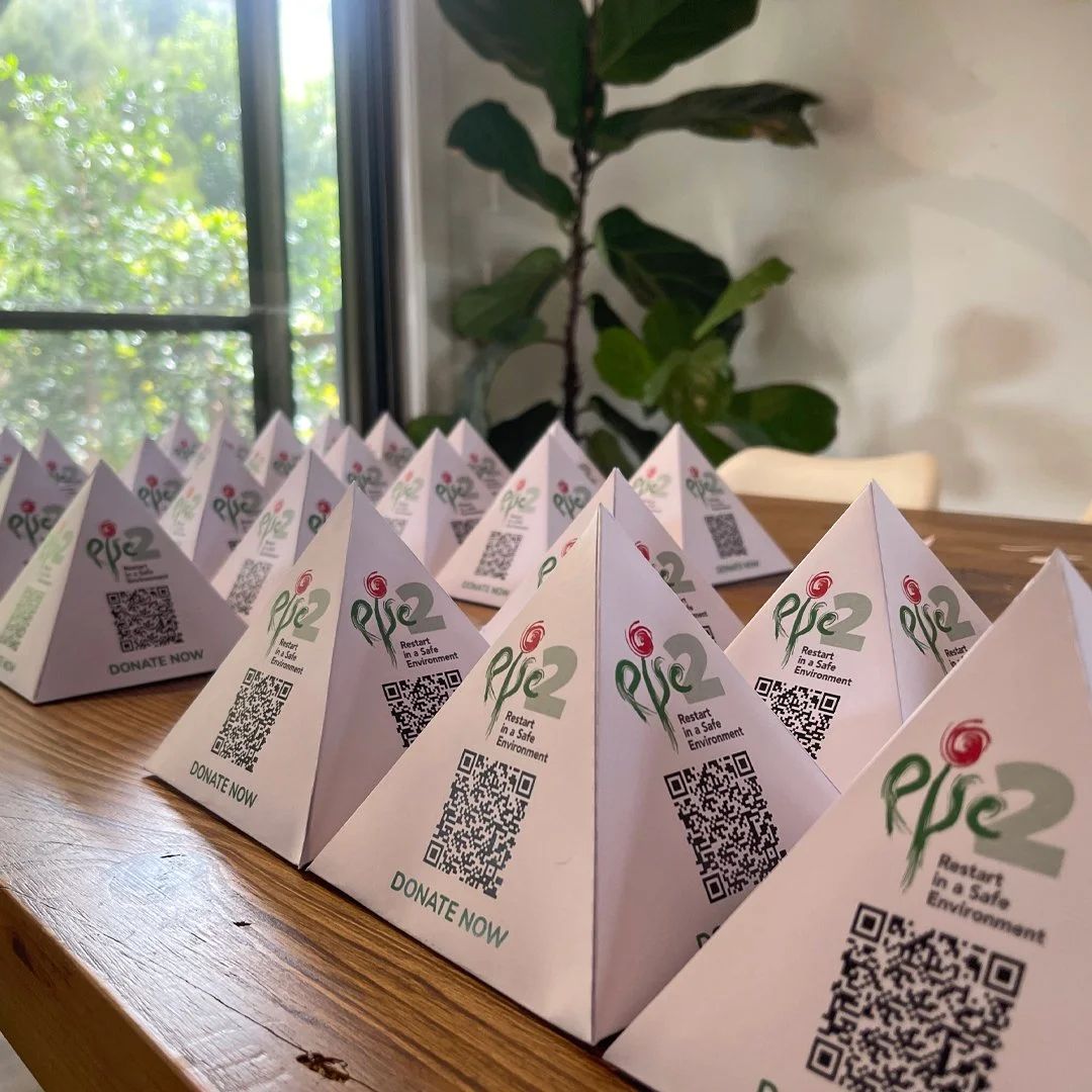 Multiple white paper pyramids with a logo, QR code, and the words 'DONATE NOW' on a wooden table near a window and a plant in the background.