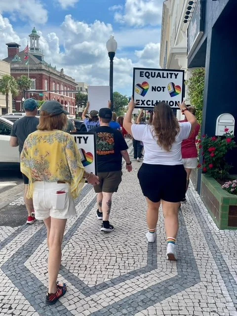 People participating in a pride parade march on a city street, holding signs advocating for equality and anti-discrimination.