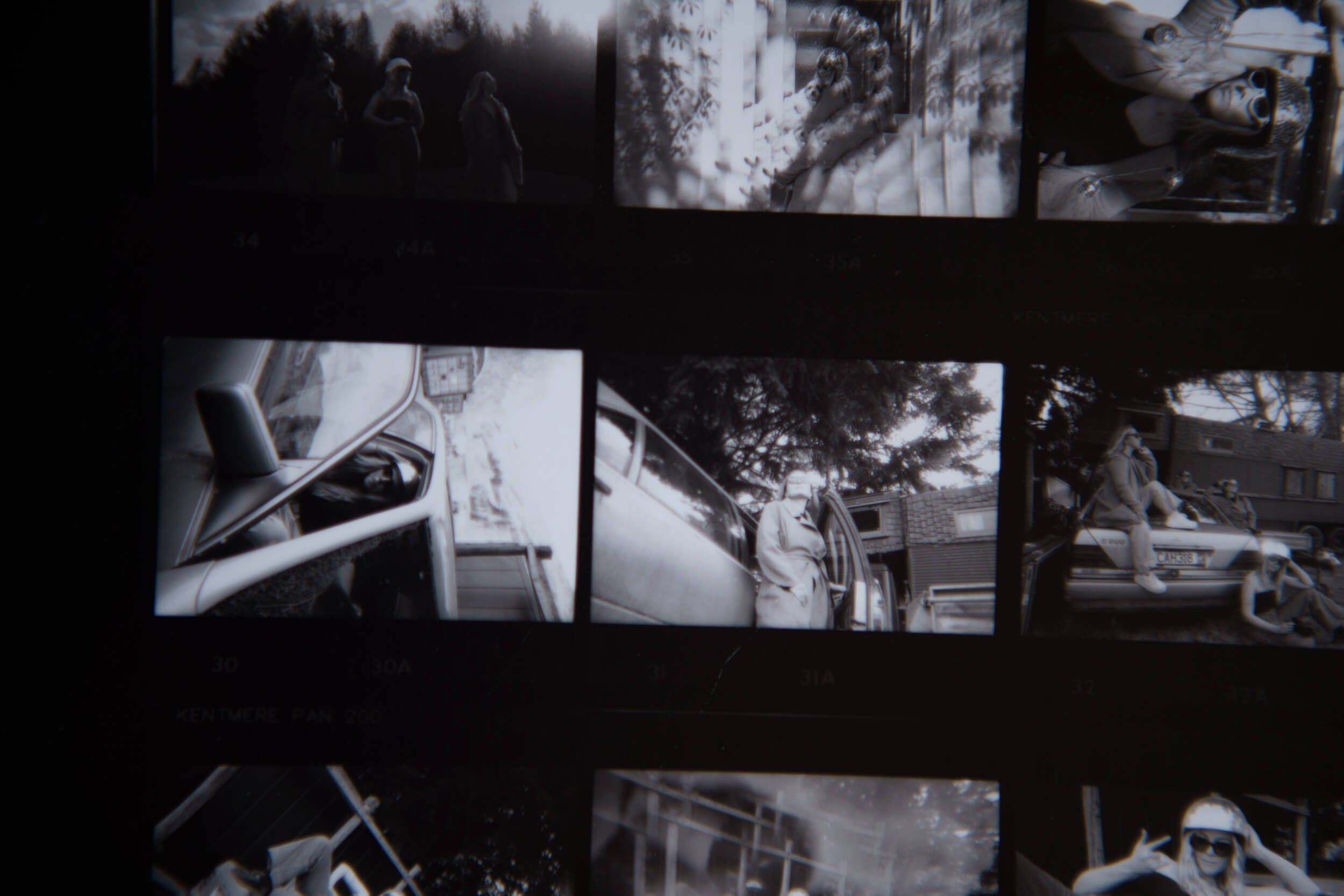 A close-up of a black and white contact sheet of multiple 35mm photographs showing various scenes, people outdoors, sitting on a bench, and around a vintage car.