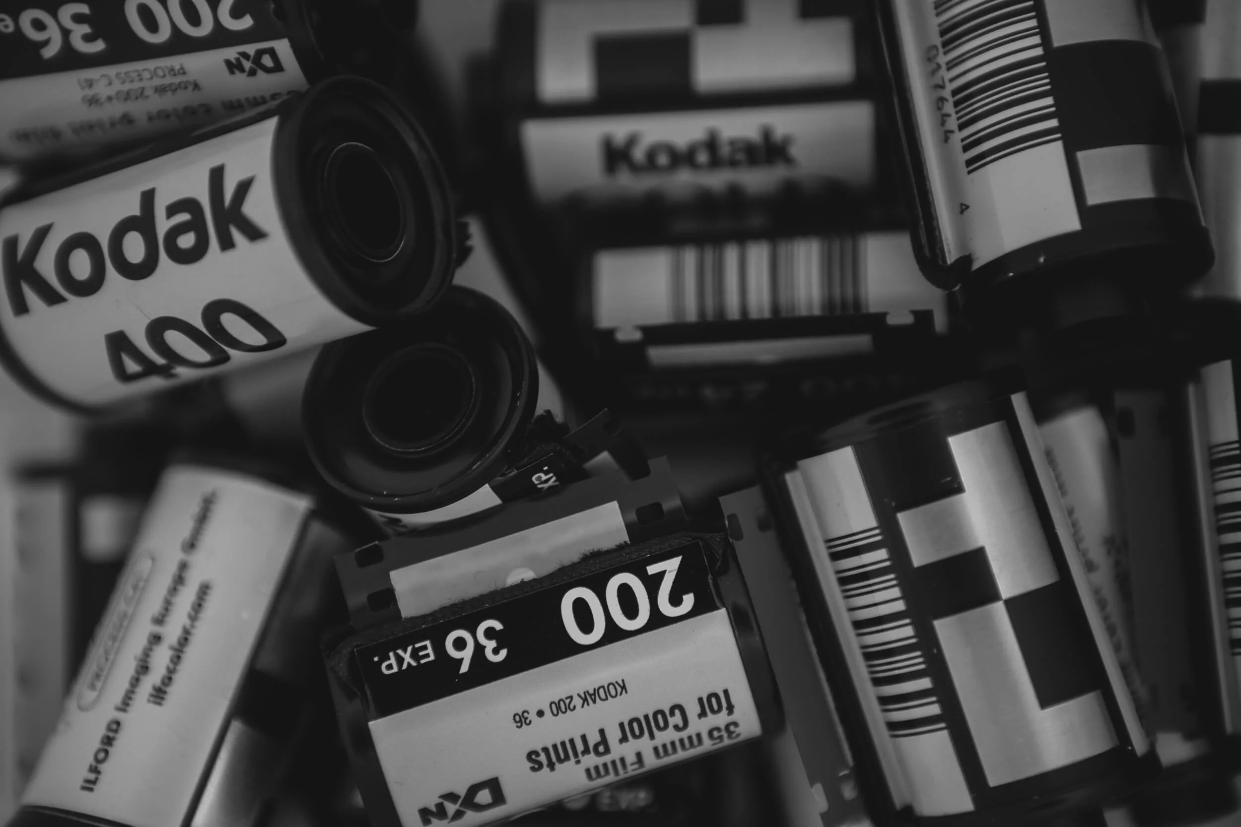 A black and white close-up of multiple Kodak 35mm colour film canisters, some with barcodes visible and labeled as 200 and 36 exposures.