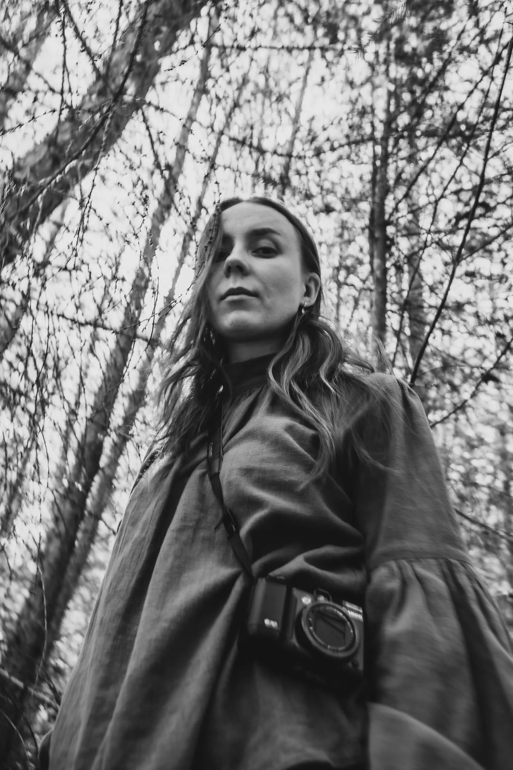 Black and white photo of a woman with long hair standing outdoors in Queenstown, surrounded by trees with bare branches, posing for and looking directly at the camera. She is wearing a retro SLR film camera on a strap around her neck.