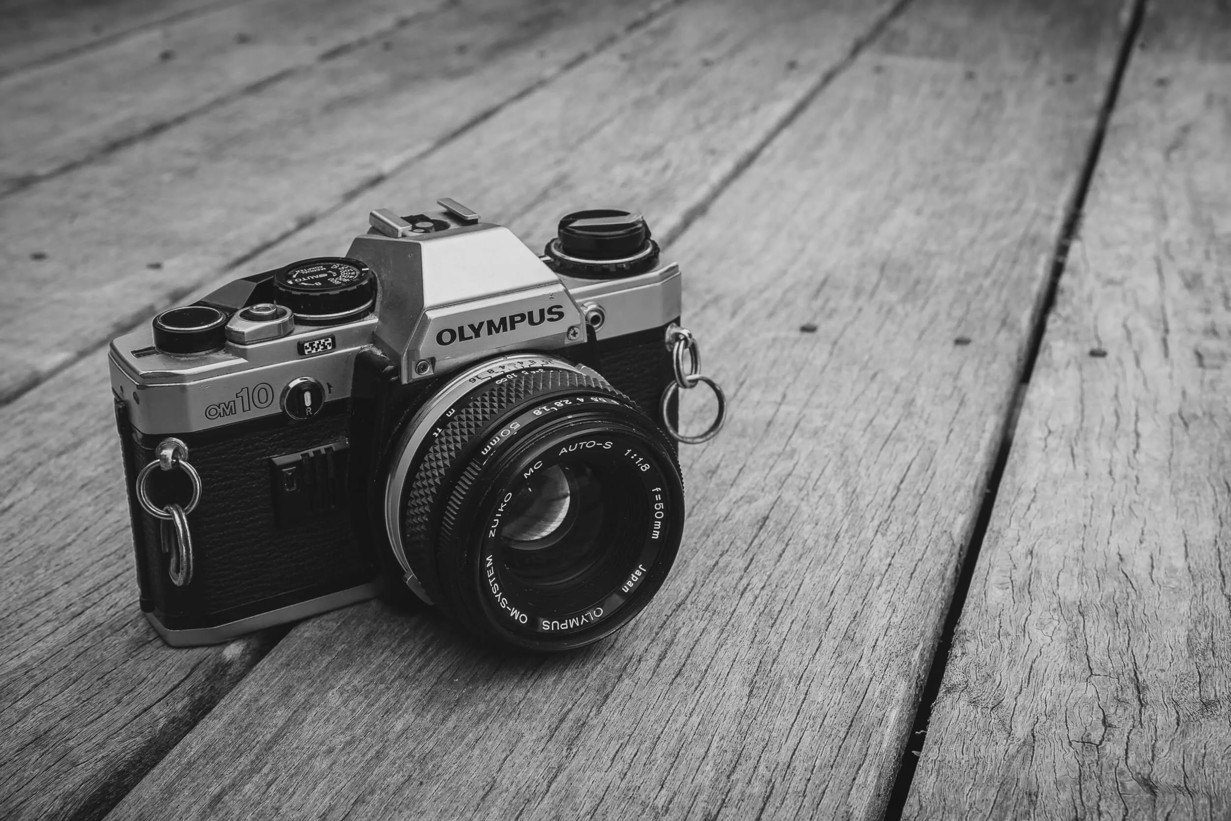 Black and white image of a classic Olympus OM-10 35mm film SLR camera with a 50mm lens on a wooden surface.