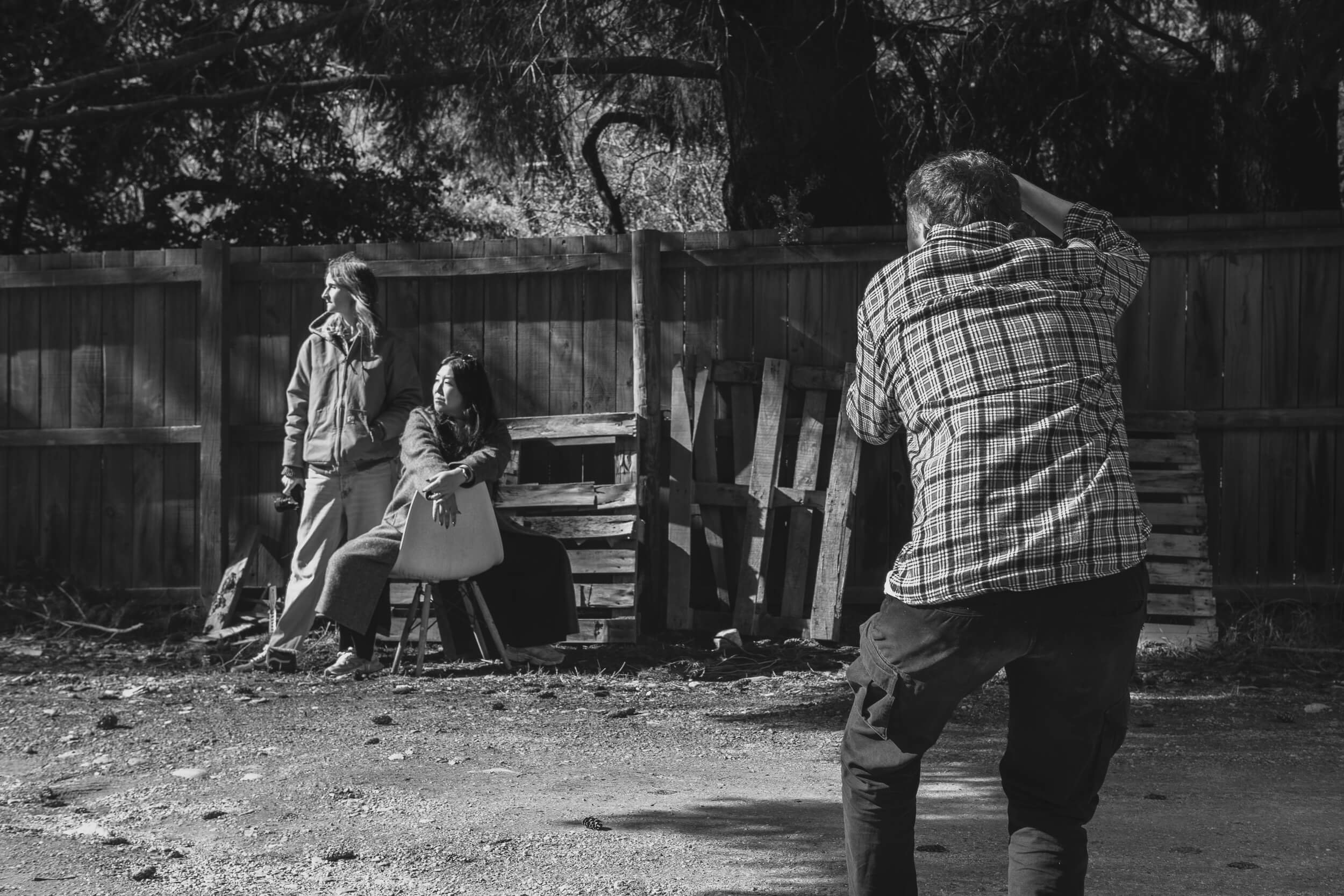 A photographer is taking a 35mm film photograph of two women posing outdoors near a wooden fence and benches; one woman is sitting on a chair, the other standing and looking to the side. The photo is in black and white.