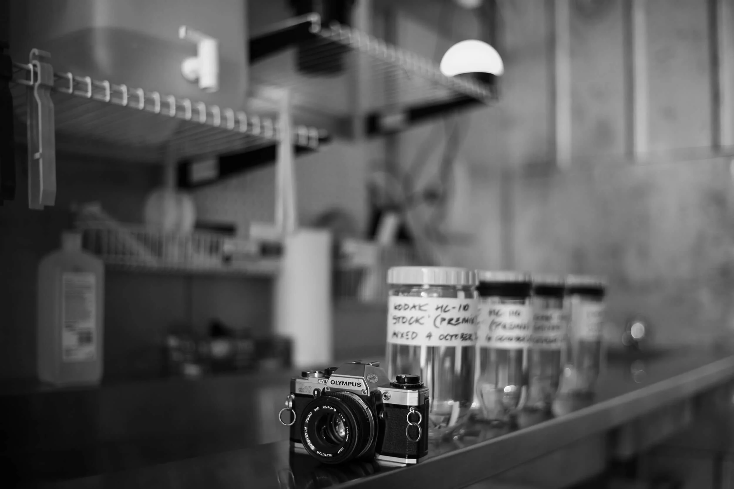 Black and white photo of a vintage Olympus camera on a stainless steel surface with glass jars full of film developer chemistry labeled with handwritten notes, in a photographic darkroom.