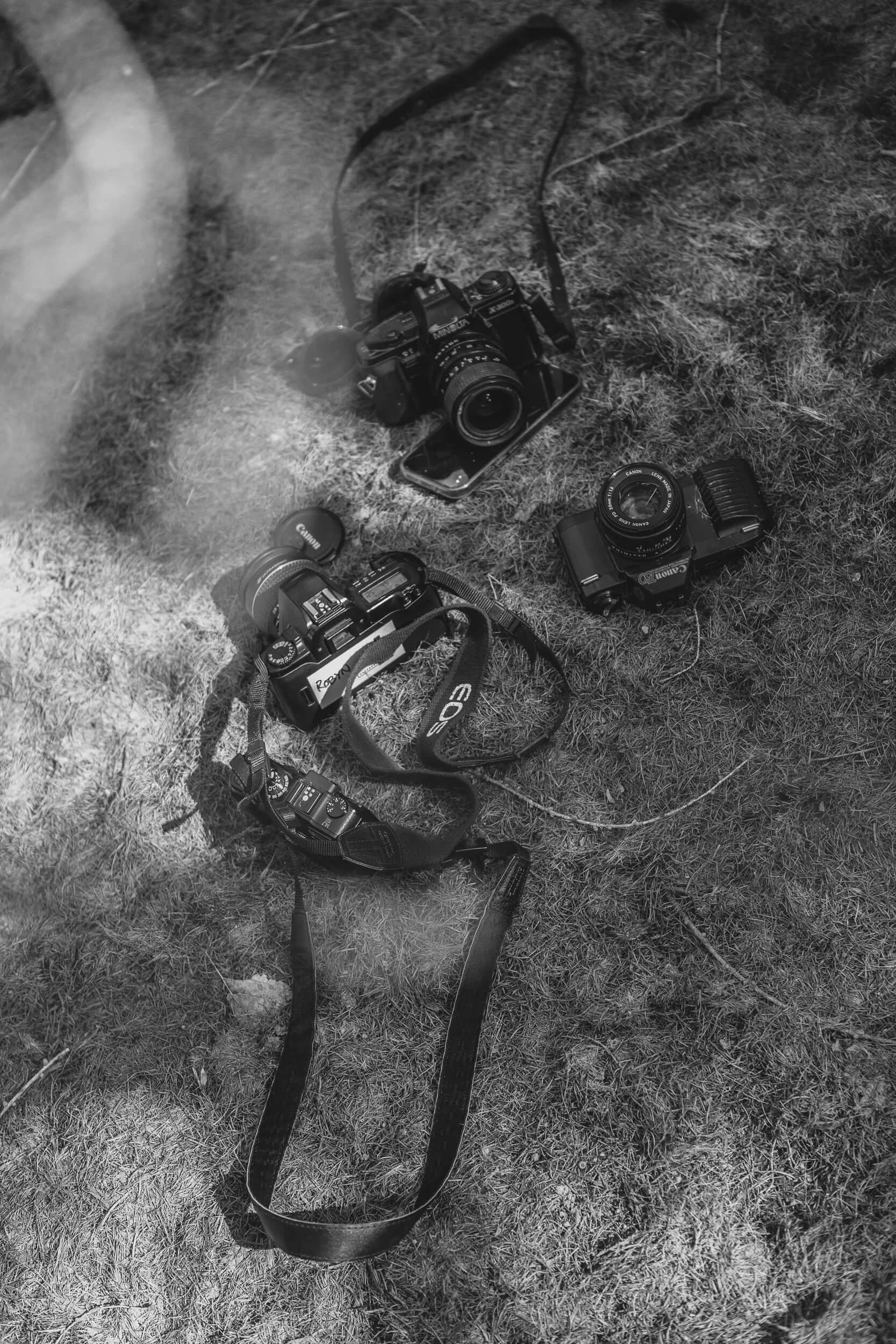 Black and White image of multiple 35mm film SLR cameras and accessories on the ground outdoors, with grass and dirt visible.