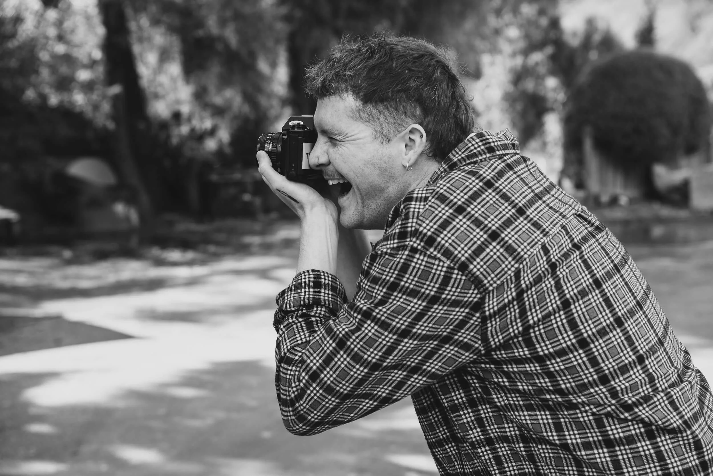 Black and White image of an enthusiastic man wearing a plaid shirt, is smiling and taking a photograph with a film camera outdoors in the Queenstown area.