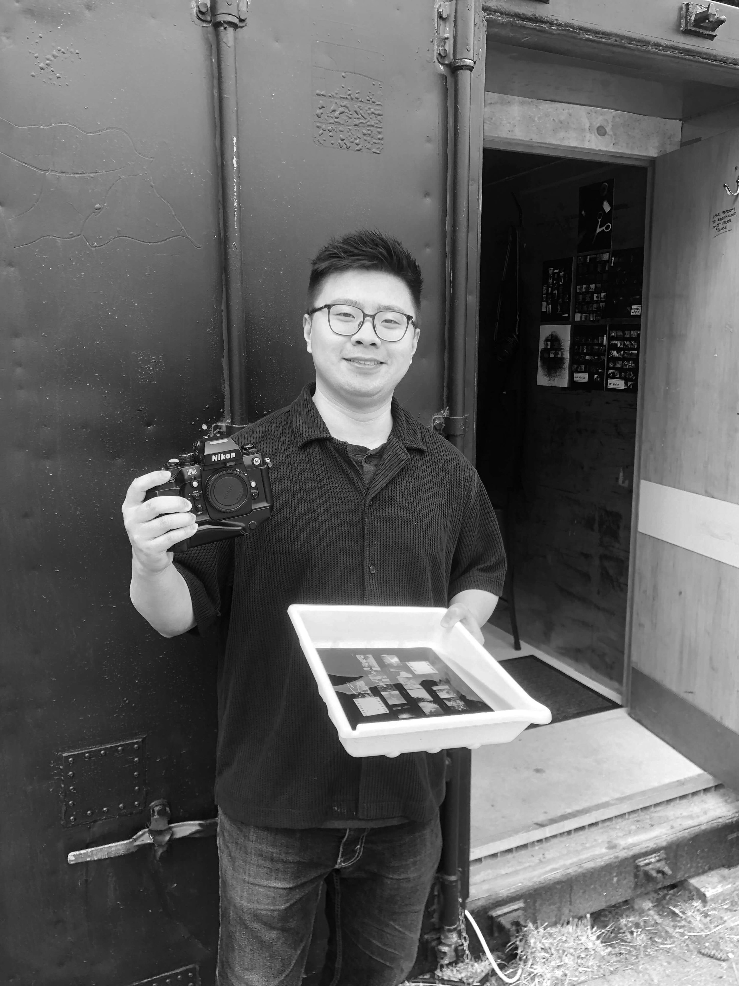 Man smiling and holding a camera and a darkroom tray while standing in front of the darkroom