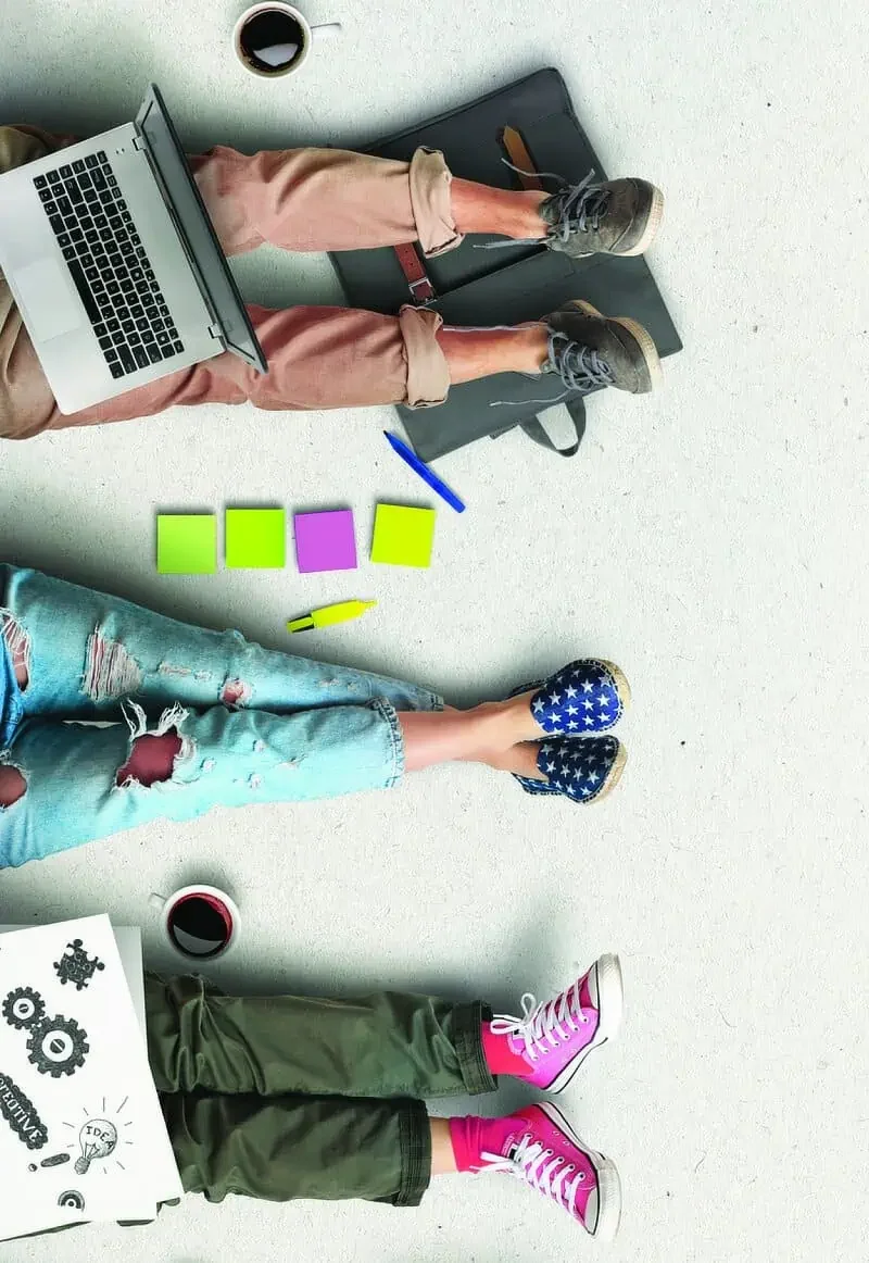 Photo of three pairs of legs stretched out, each wearing colourful sneakers and casual clothing. A laptop, cups of coffee, and colourful sticky notes are visible on the floor.