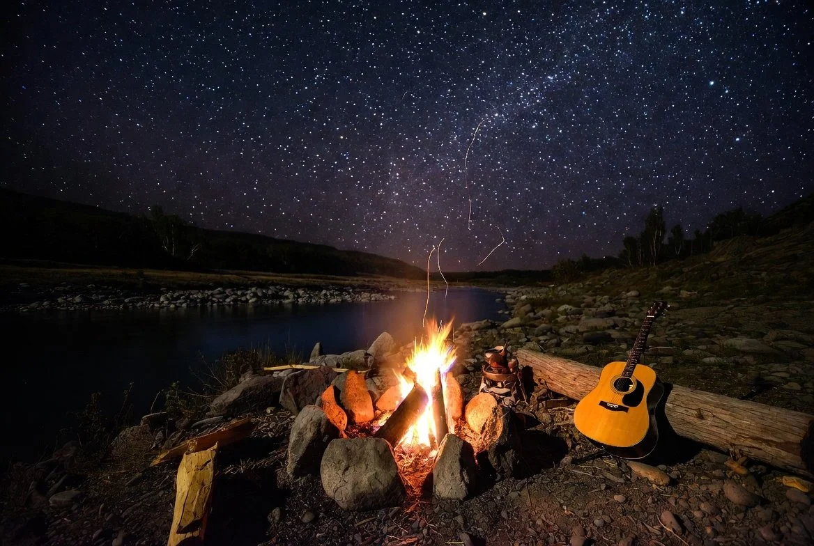 Night scene by a river with a campfire, guitar, and trees in the distance under a starry sky.