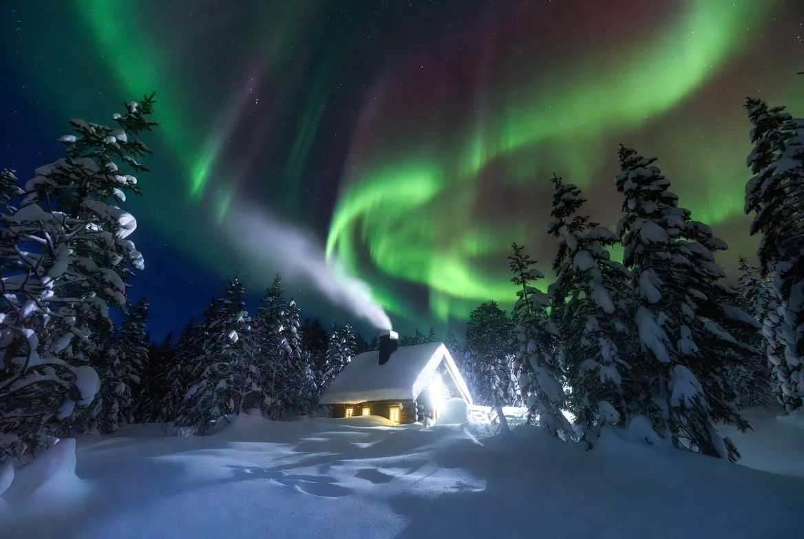 A snowy landscape at night with a cabin emitting smoke from its chimney, surrounded by snow-covered trees, under a sky illuminated by the colorful northern lights (aurora borealis).