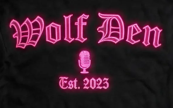Pink neon sign with the words 'Wolf Den' in Gothic font above a microphone icon and 'Est. 2023' below, on a dark background.