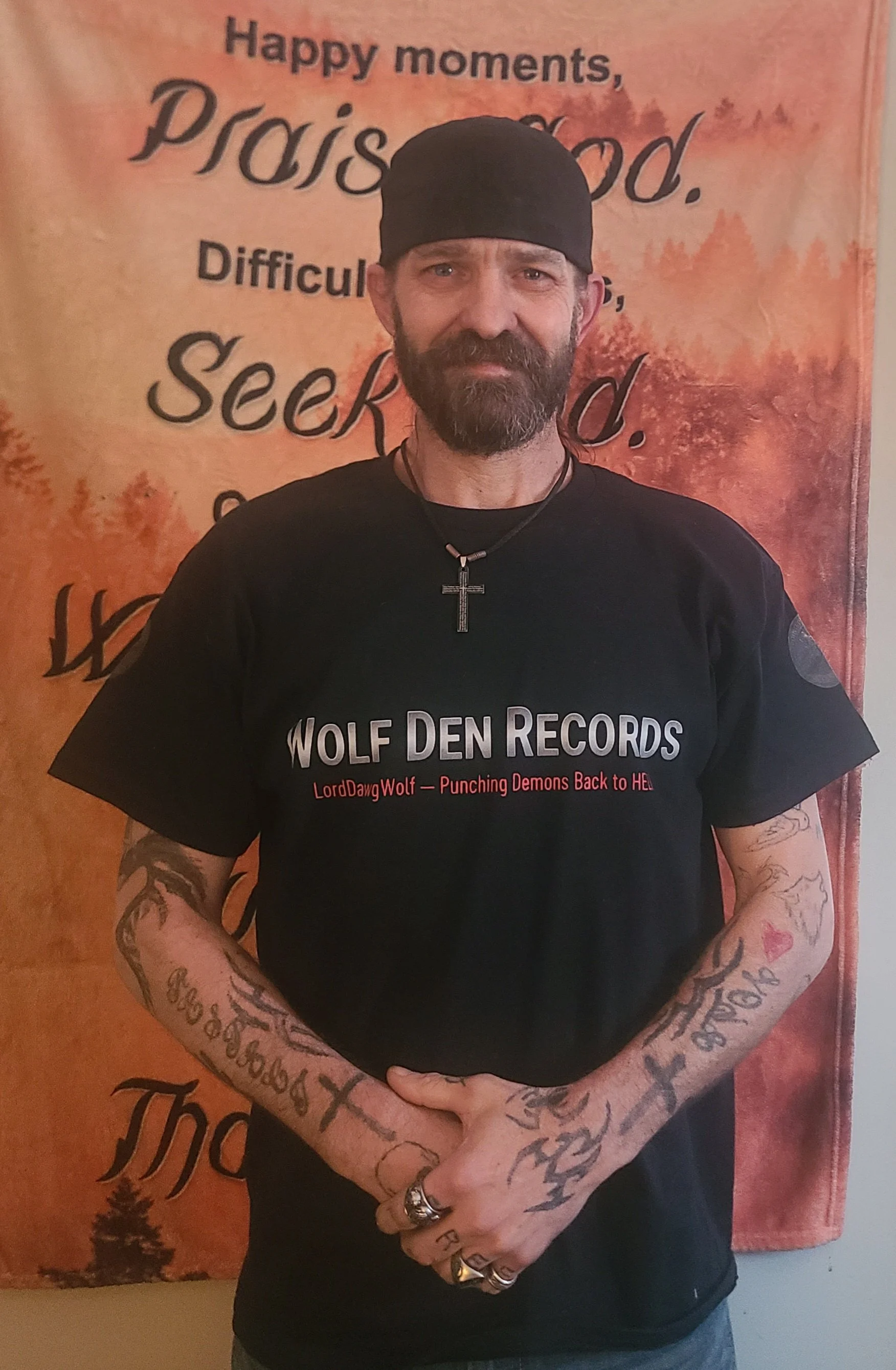 Man with a beard and tattoos wearing a black beanie and black Wolf Den Records t-shirt, standing in front of a burnt orange background with black text and trees, smiling.