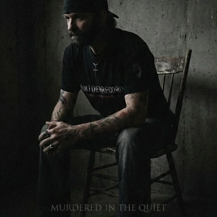 A man with a beard and tattoos, wearing a black t-shirt and cap, sitting on a wooden chair in a dimly lit room with concrete walls, looking pensively downward.