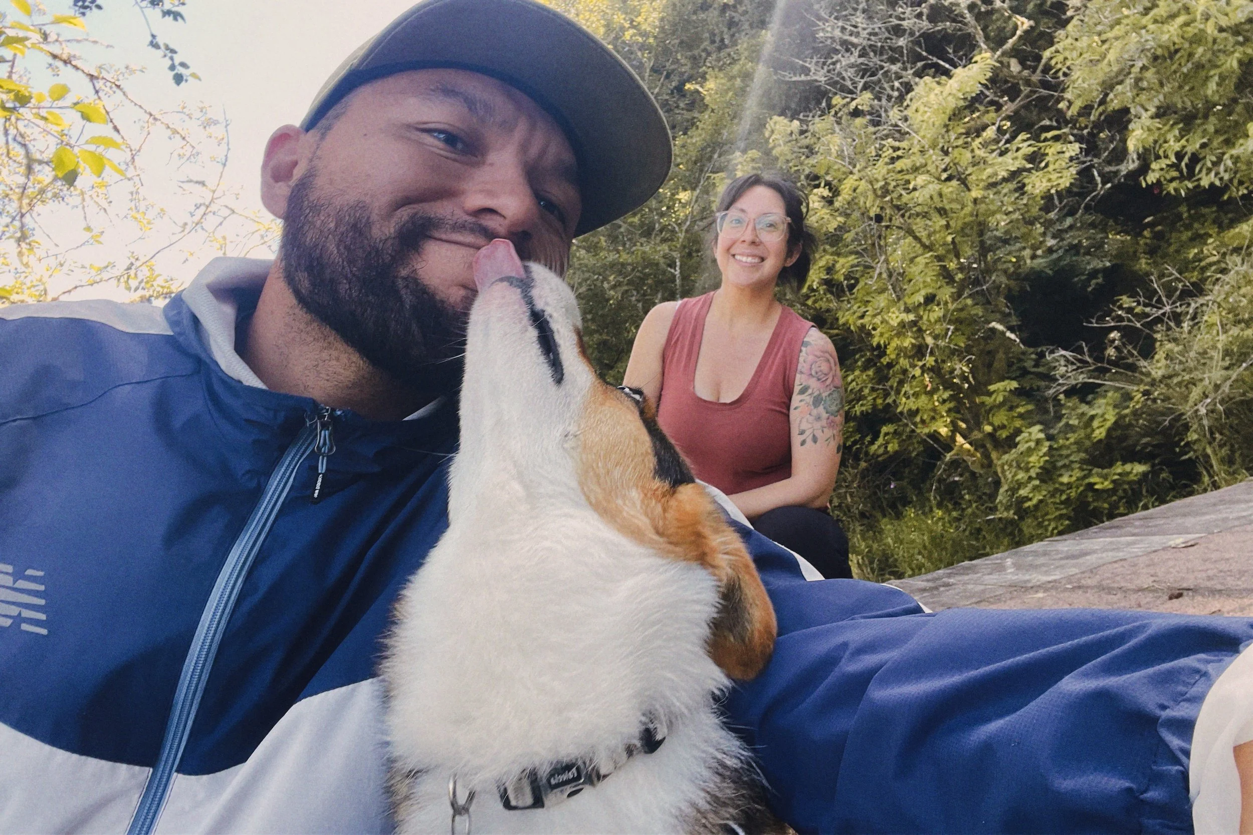 Man with beard wearing a blue jacket and cap, dog licking his face, woman with tattoo on arm smiling in the background, outdoors with trees.