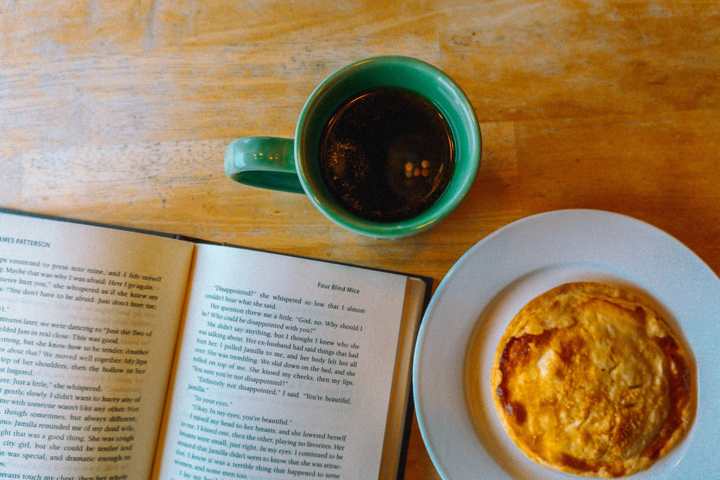 Top-down view of a green mug filled with black coffee on a wooden table, partially visible a book and a plate with a baked dish.