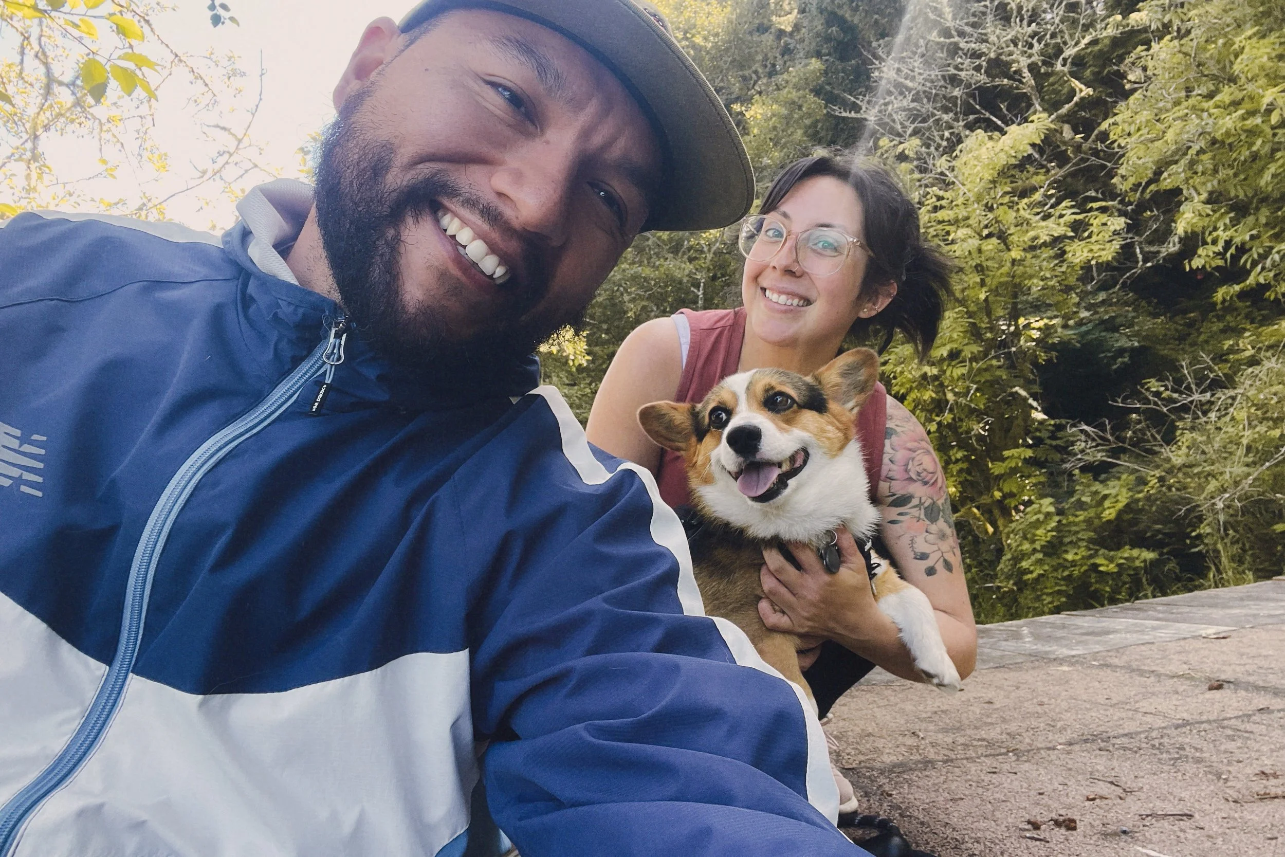 A man and woman smiling with a joyful dog in an outdoor setting with green trees in the background.
