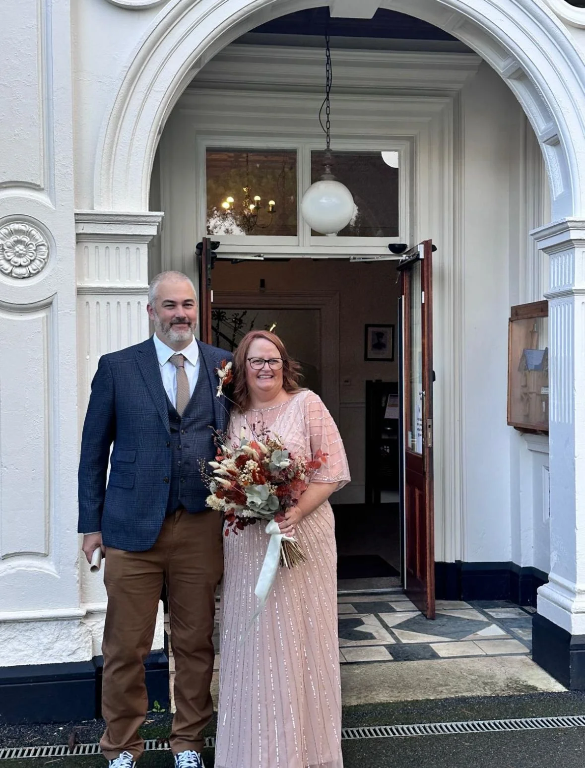 A man in a navy blazer and a woman in a peach-colored beaded dress holding a large autumnal-themed dried flower bouquet.