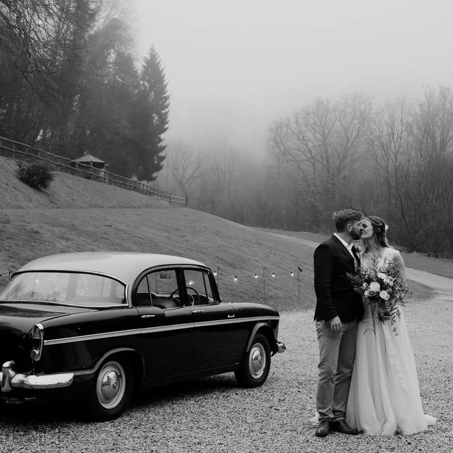 A black and white photo of a couple in wedding attire kissing outdoors next to a vintage car on a gravel road with a misty, wooded hillside in the background.