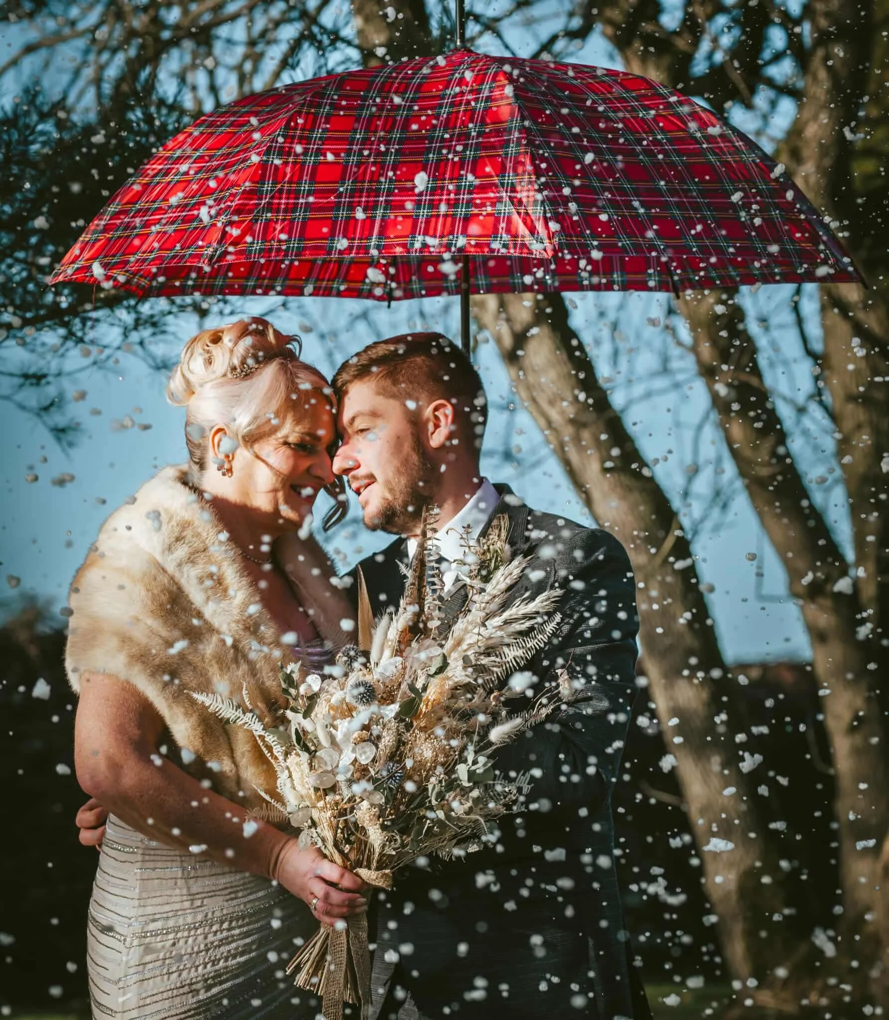 A newly married couple, the bride in a fur stole and the groom in a suit, embrace under a red plaid umbrella while confetti falls around them.