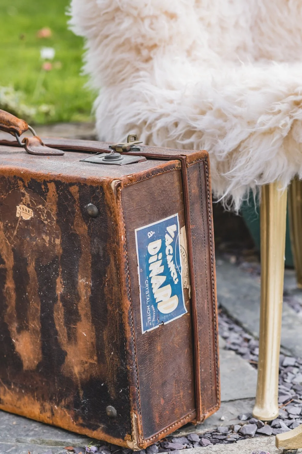 an old vintage suitcase on the ground next to a gold chair with a sheepskin fur rug draped over it. Symbolizing waiting to go on an adventure