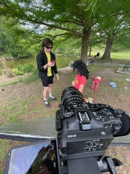 A woman with short black hair and sunglasses holding a smartphone stands next to a girl with curly black hair, wearing a red shirt with a yellow logo, under a large green tree near a pond.Recording equipment is set up in the foreground, pointing towa