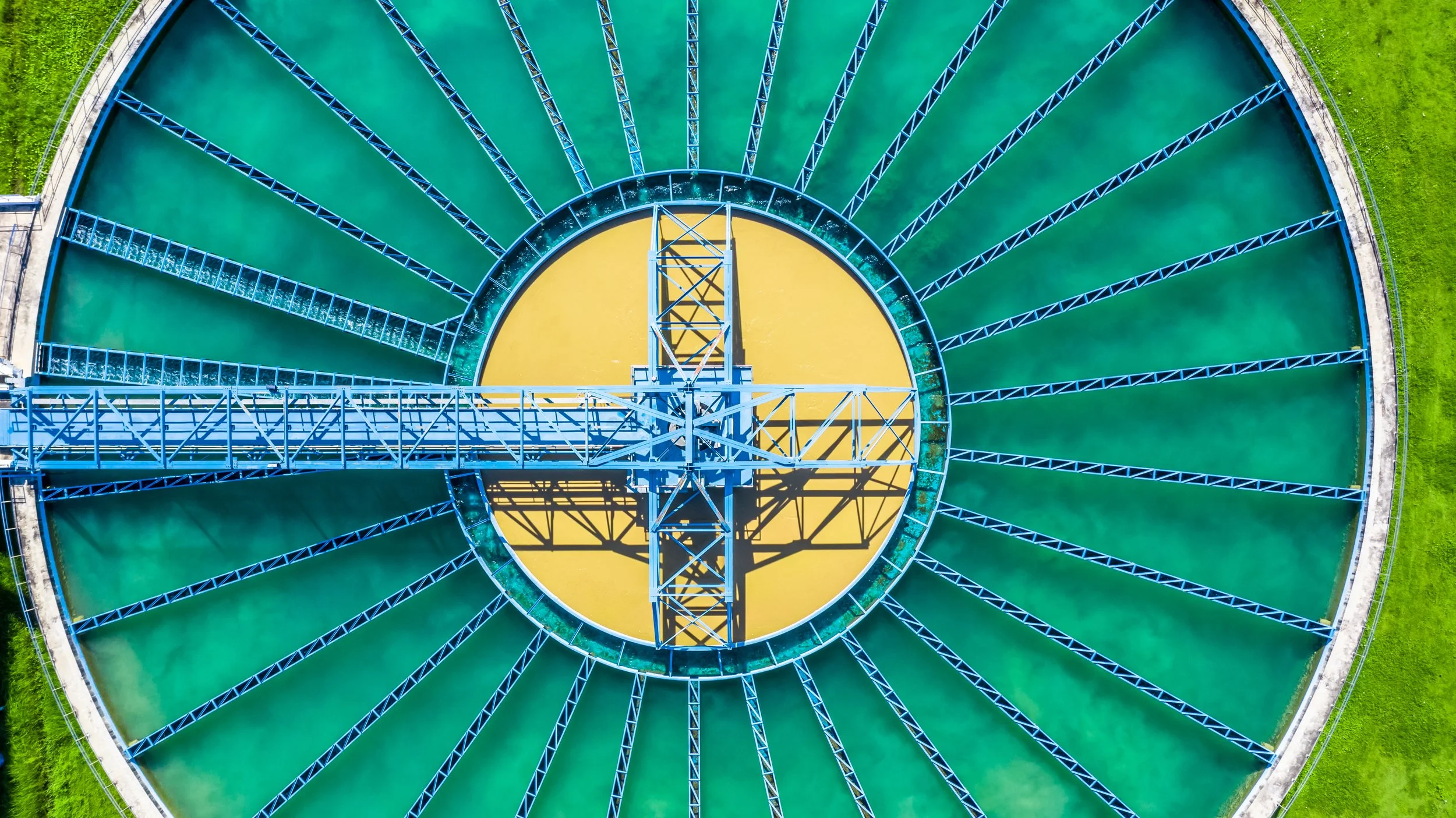 An aerial view of a circular water treatment tank with a yellow bottom, green water, and a metal walkway crossing the center.