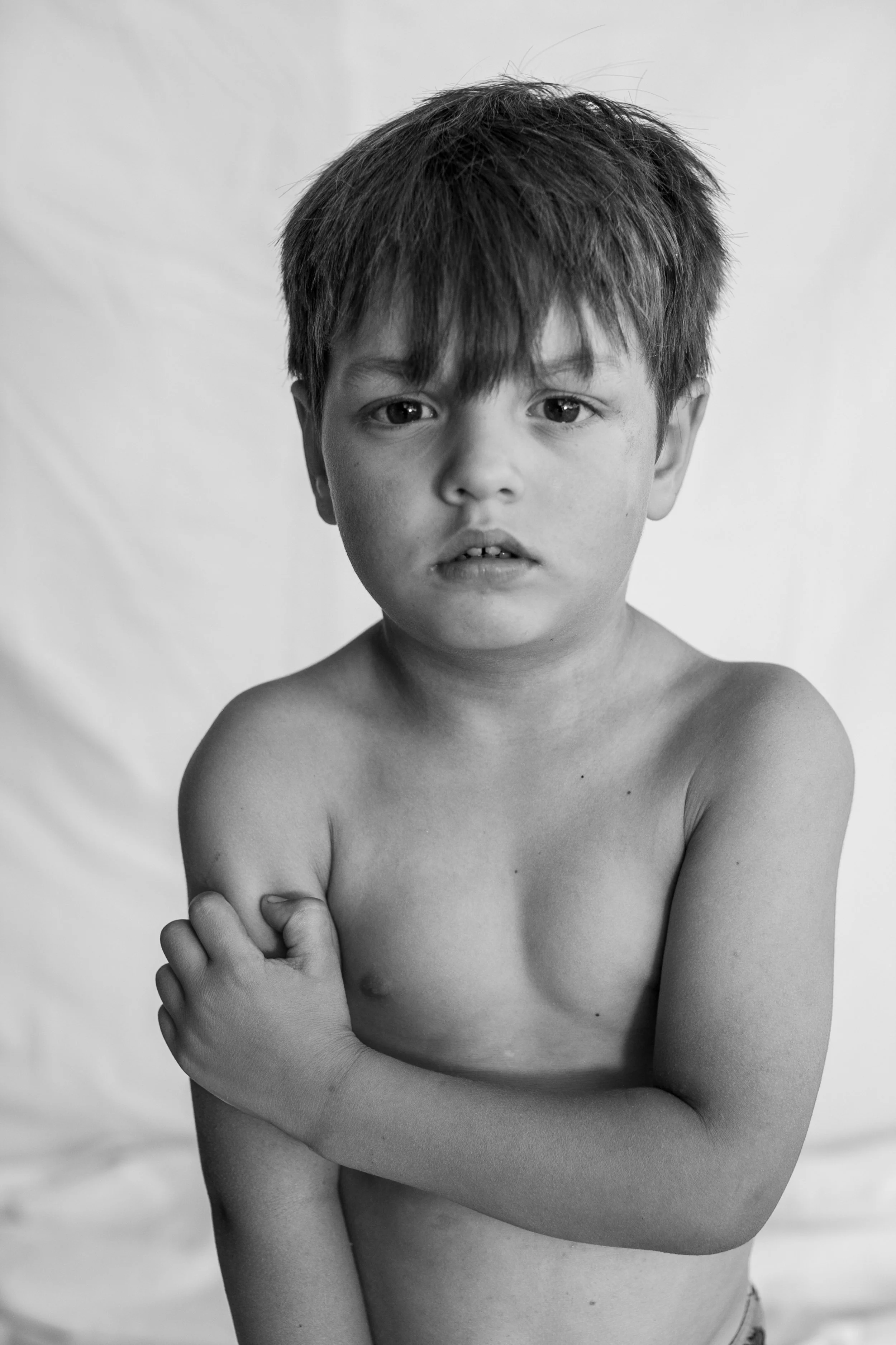 A young shirtless boy with tousled hair and a serious expression, holding his left arm with his right hand. The background is plain and light-colored.