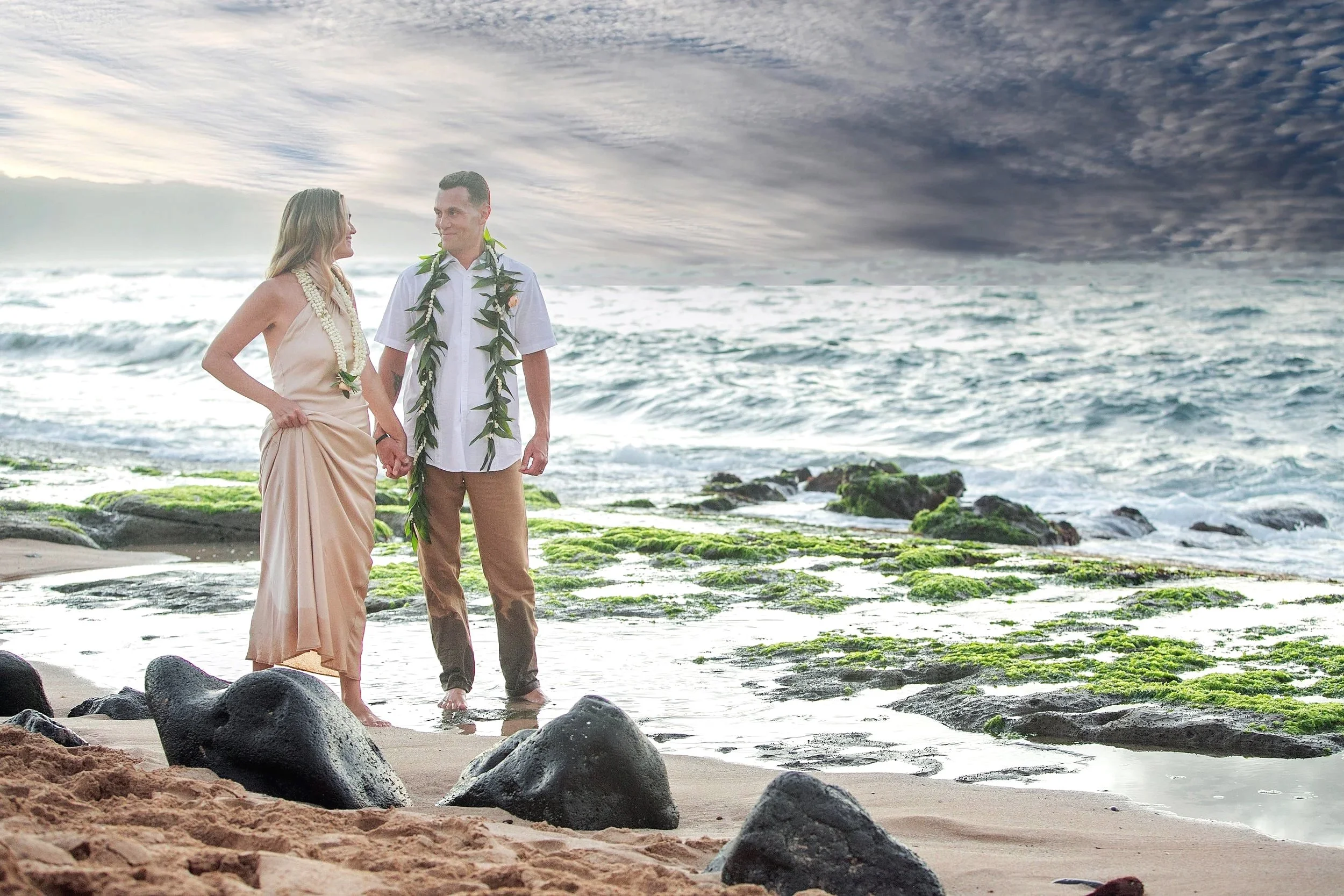 A couple standing hand in hand on a rocky beach, dressed in traditional Hawaiian wedding attire, with green moss on rocks and an ocean backdrop under a cloudy sky.