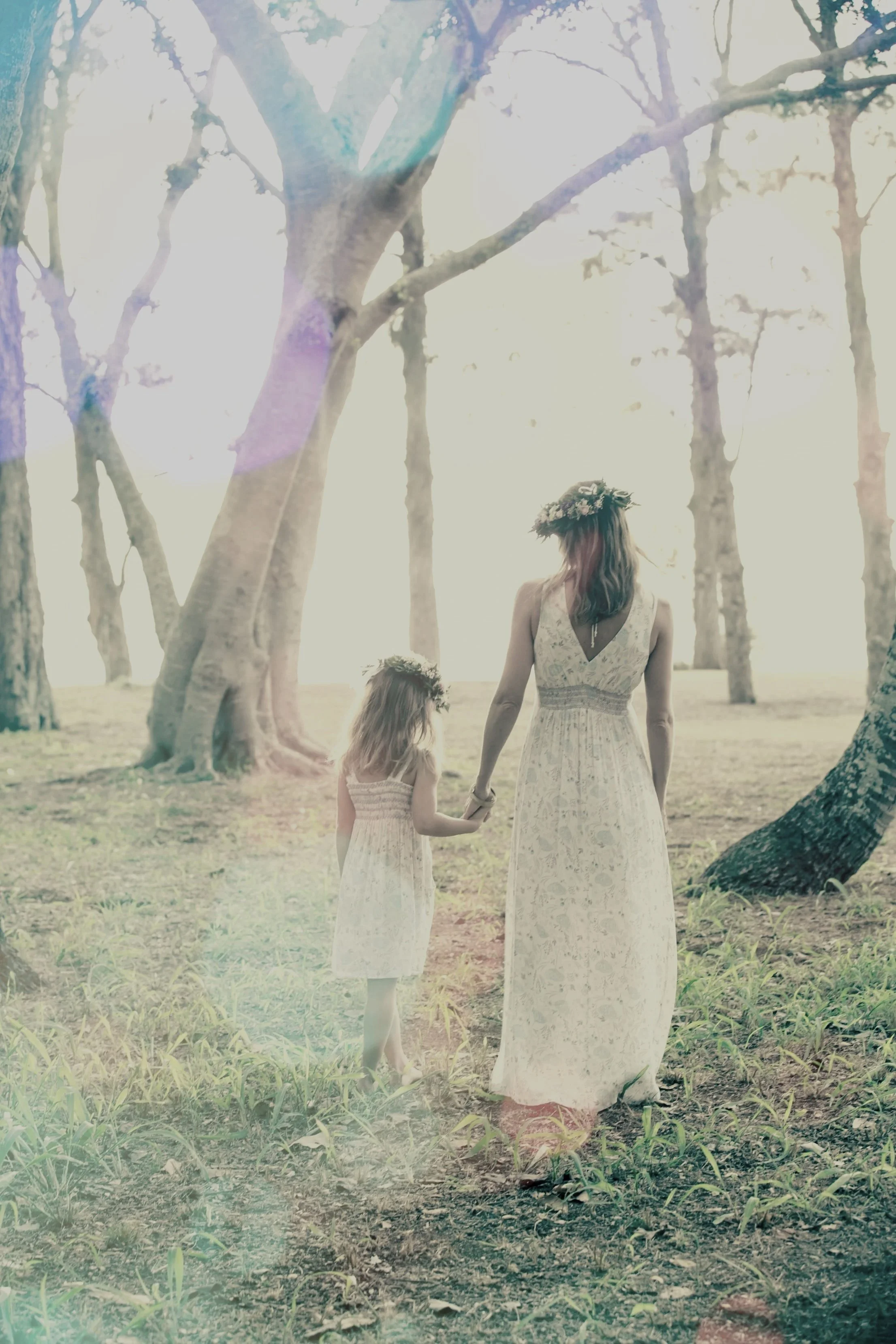 A woman and a young girl, both dressed in white dresses, walking hand in hand through a sunlit forest with tall trees, wearing flower crowns.
