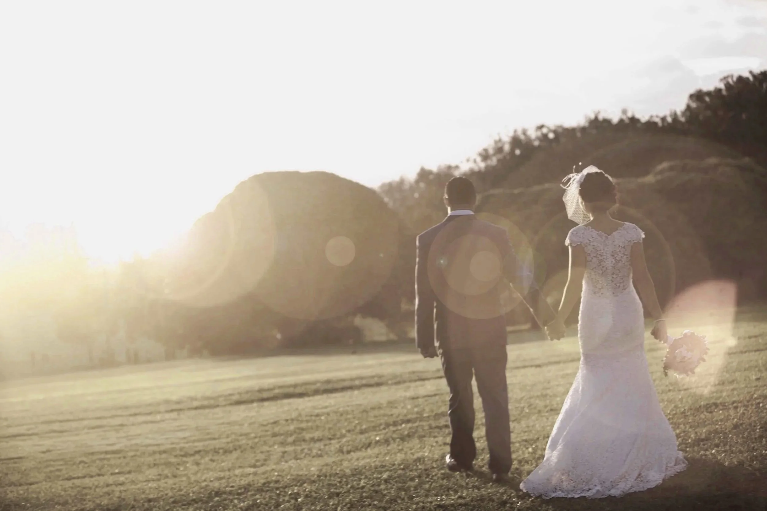 A bride and groom walking hand in hand on a grassy field during sunset, with the bride holding a bouquet, dressed in a wedding gown and veil, and the groom in a suit.