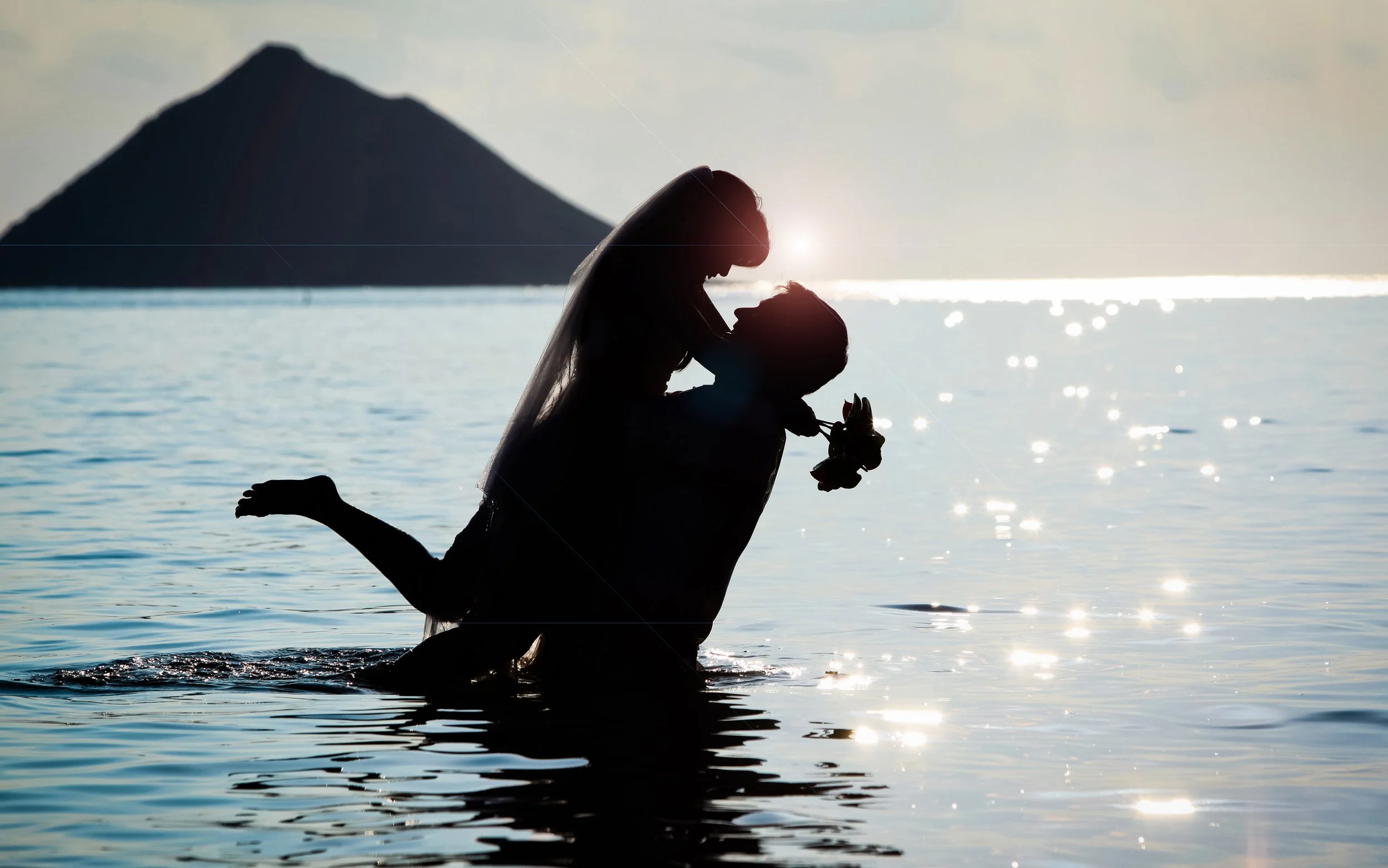 Silhouette of a bride holding a bouquet, kneeling in water during sunset, with an island and mountain in the background.