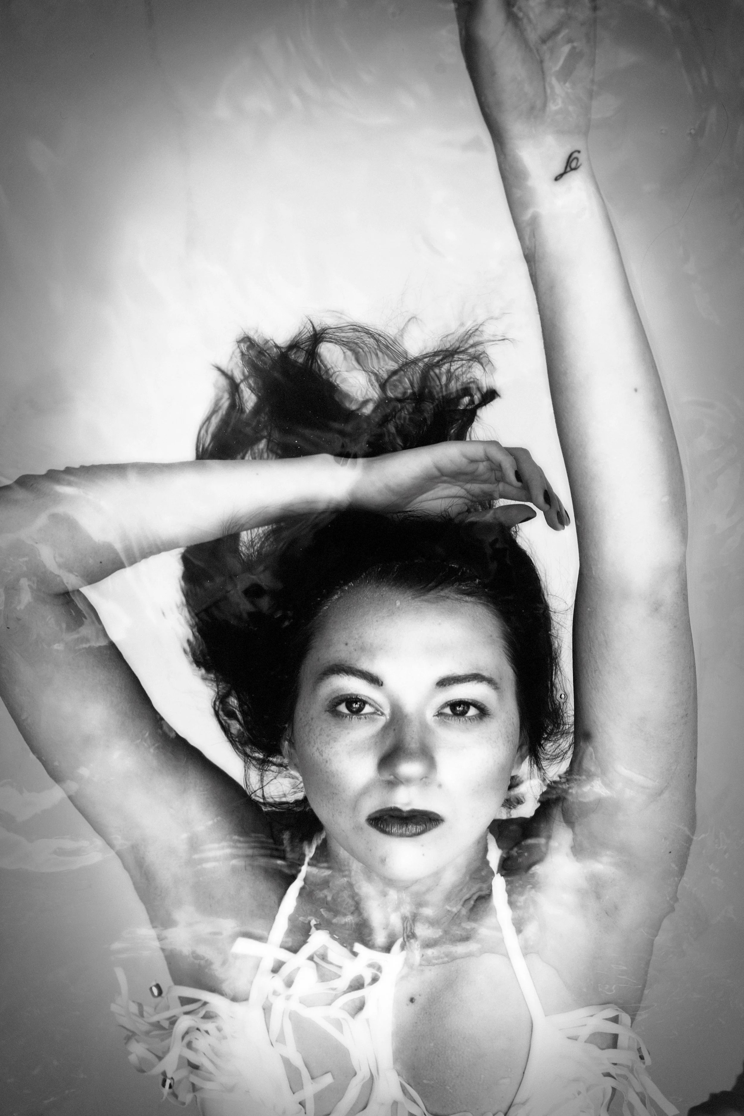 A black and white underwater photograph of a woman with dark hair and lipstick, lying in the water with her arms above her head and a small tattoo on her right forearm.