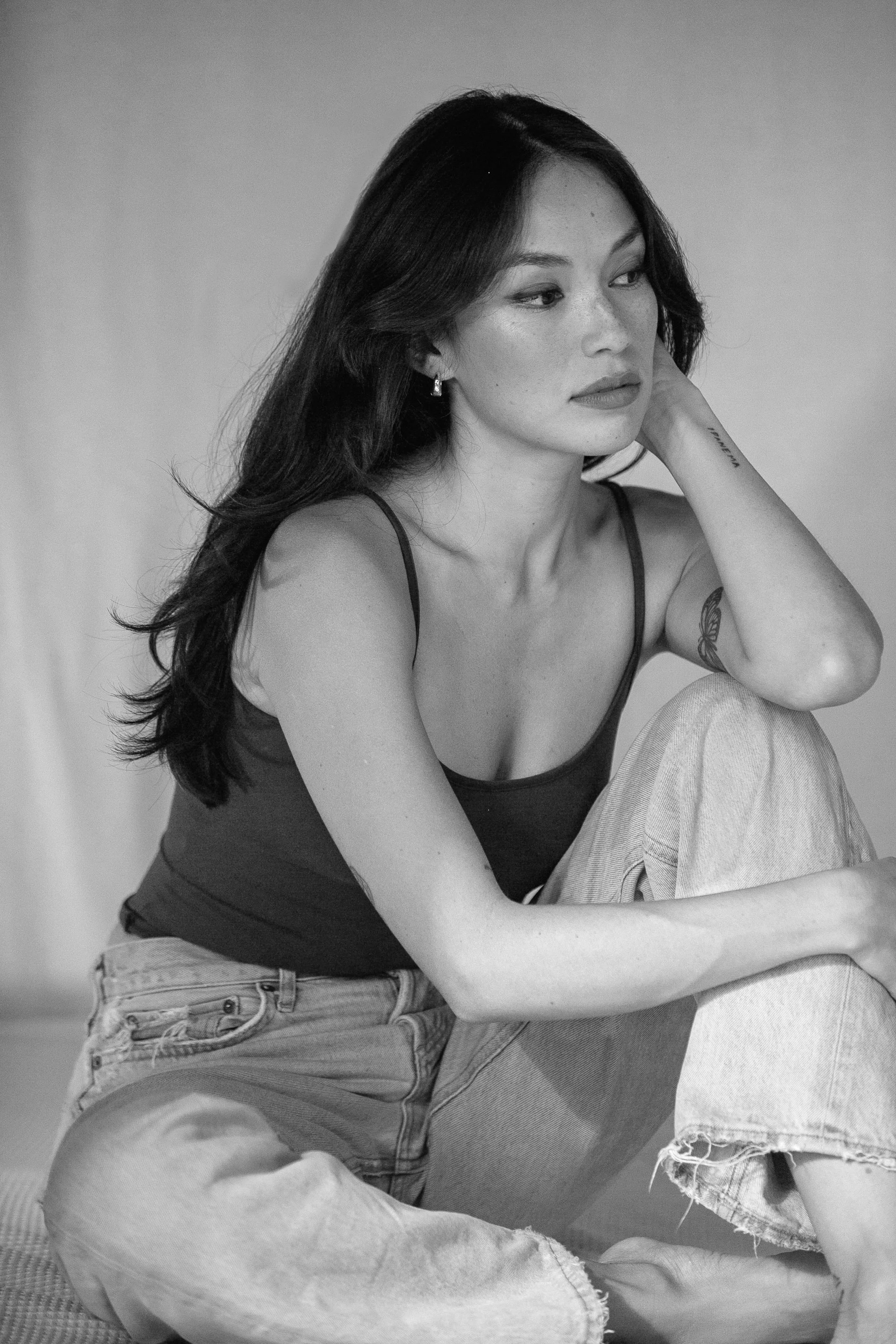 Black and white photo of a young woman with long dark hair, wearing a tank top and ripped jeans, sitting with one knee up and resting her face on her hand, looking thoughtfully to the side.