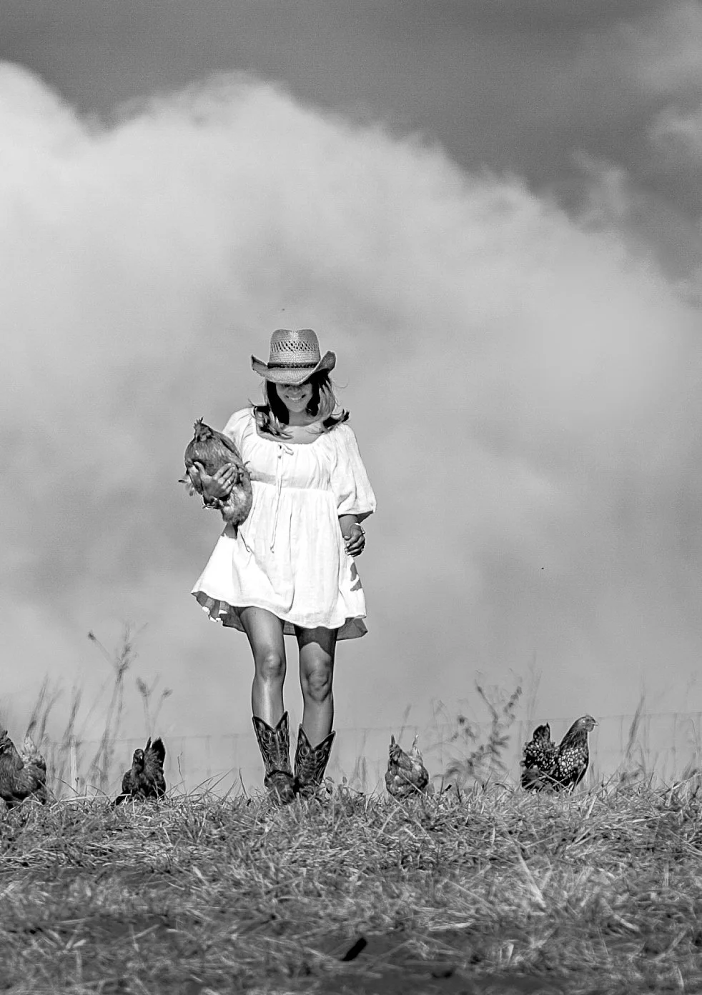 A girl in a dress and cowboy boots walking on a grassy field holding a chicken, surrounded by other chickens, with a sky and clouds in the background. She is wearing a wide-brimmed hat and smiling.