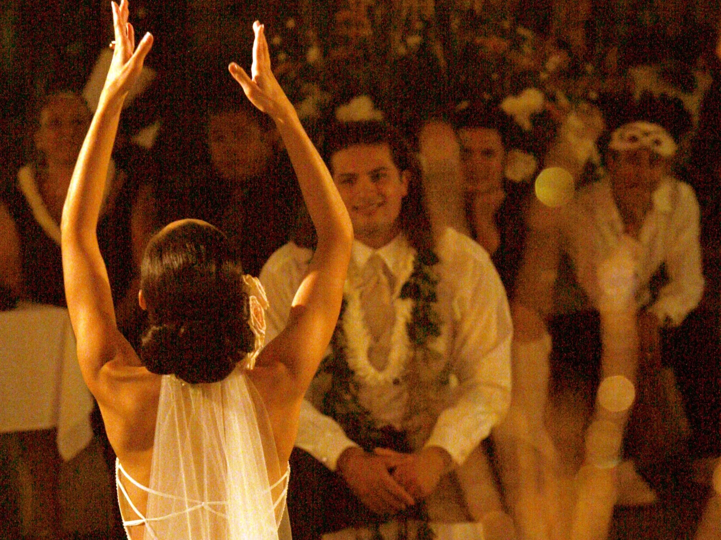A young girl dressed as an angel, with a veil and a golden hair clip, is performing a dance with her arms raised. She is facing a seated man in a white shirt and lei, who is smiling and watching her. Several other people are standing in the background, observing the performance.
